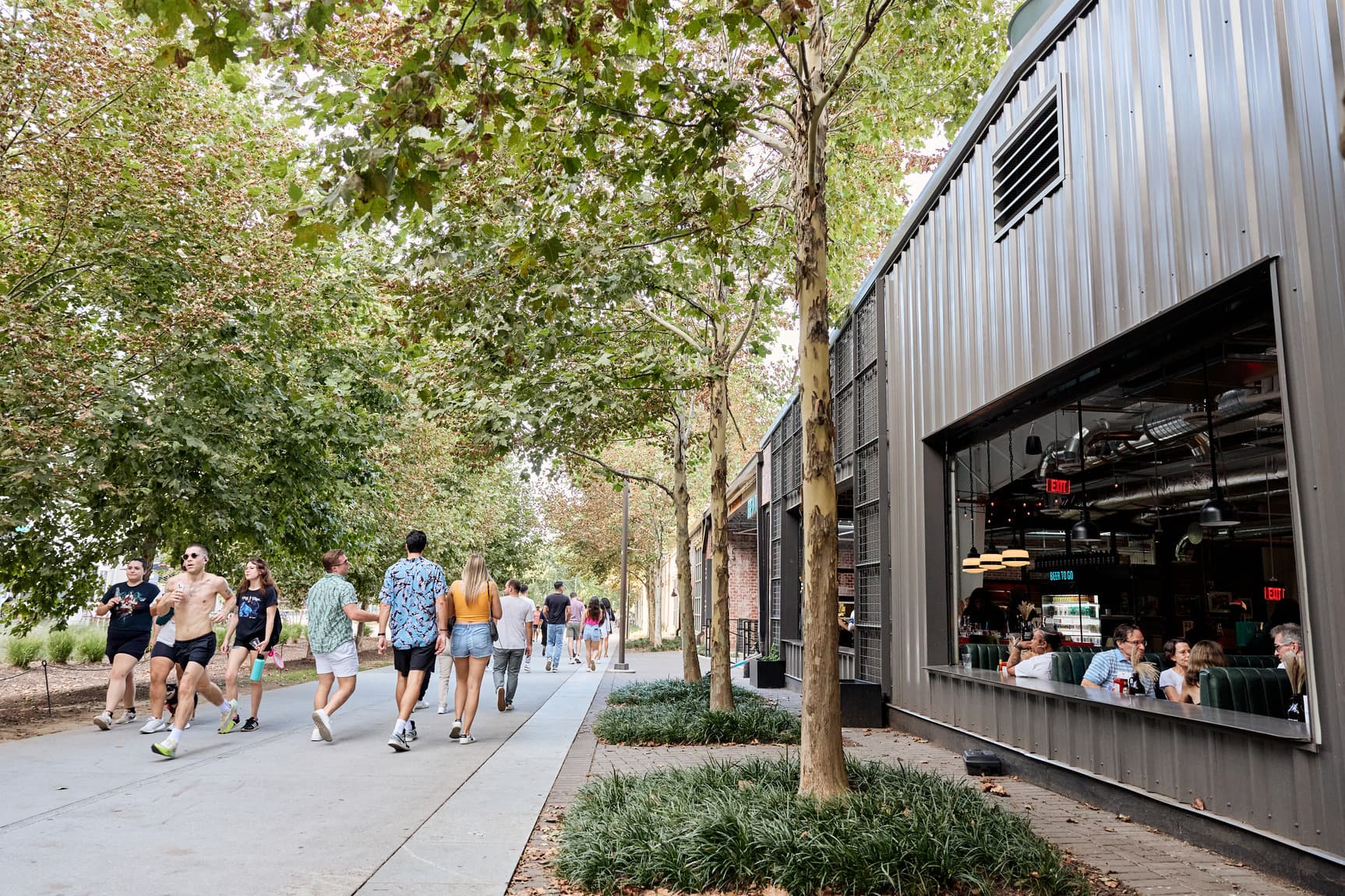 A group of people walk along the Eastside Trail. Trees line either side of the path and to the right is a large restaurant with an open air space.